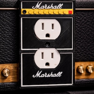 May include: Black and white electrical outlet cover with the word "Marshall" in white script. The top section has a yellow and red design. The outlets are white. The cover is mounted on a black surface.