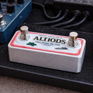 May include: A silver guitar effects pedal with the text "ALTIODS" in a red and white label. The pedal has two silver foot switches and is surrounded by other guitar effects pedals on a black surface.