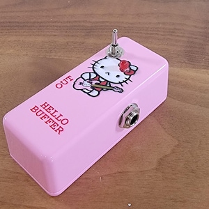 May include: A rectangular pink guitar buffer pedal featuring a graphic of Hello Kitty playing a guitar. The pedal includes a metal toggle switch, a red LED indicator light, and a side-mounted input jack. The text HELLO BUFFER and OUT are printed on the top surface in red.