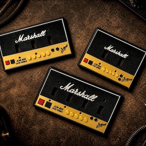 May include: Three black and gold Marshall JCM 800 Lead Series cassette tape-shaped keychains. Each keychain has the Marshall logo in white, with a gold and black design. The keychains are on a brown leather surface.