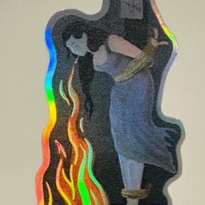 Puede incluir: Pegatina holográfica que presenta una ilustración estilizada de una figura con un vestido azul, atada y de pie entre llamas. Las llamas son vibrantes con tonos naranja, amarillo y verde. La figura está frente a un fondo oscuro.