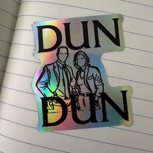 May include: Holographic sticker featuring a stylized illustration of two people, with the word "DUN" above and below the figures. The figures are in black outline, and the sticker has a rainbow sheen.