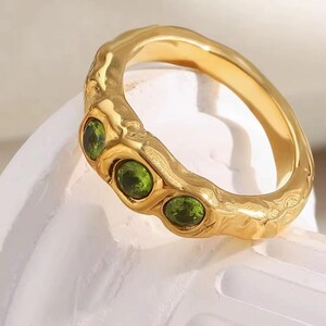 Könnte beinhalten: Ein goldfarbener Ring mit einem strukturierten Band und drei ovalen, grünen Edelsteinen. Der Ring ist vor einem weißen Hintergrund platziert, was seine polierte Oberfläche und die lebendige Farbe der Steine hervorhebt.