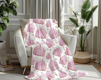 Puede incluir: Una manta blanca con un patrón repetido de casas de jengibre rosas, lazos y detalles dorados. La manta está sobre un sillón blanco. El diseño incluye estrellas y detalles decorativos.