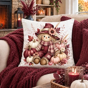 May include: A square throw pillow featuring a central illustration of a scarecrow wearing a maroon hat and scarf, surrounded by various pumpkins in shades of white, cream, and maroon, accented with autumn leaves, sunflowers, and wheat stalks.