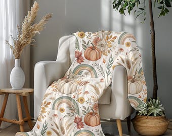 May include: A cream-colored throw blanket draped over a white armchair. The blanket features a fall-themed pattern with pumpkins, rainbows, sunflowers, and foliage in shades of orange, brown, and green. The blanket is made of a soft material.