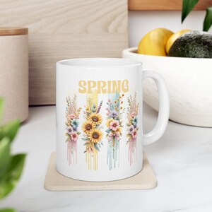May include: White ceramic mug with a floral design and the word "SPRING" in yellow letters. The flowers are in shades of pink, yellow, blue, and green. The mug has a handle on the right side.