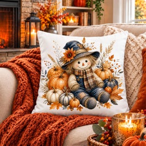 Peut inclure: Un coussin carré blanc orné d'une illustration centrale représentant un épouvantail souriant portant un chapeau bleu foncé avec un tournesol, une écharpe à carreaux et une salopette bleue, entouré de citrouilles orange, blanches et bleues.