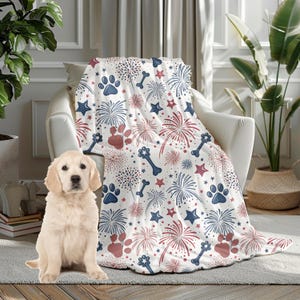 May include: A white throw blanket with a pattern of red and blue fireworks, paw prints, stars, and dog bones. The blanket is draped over a white armchair. A golden retriever puppy sits on the floor in front of the chair.