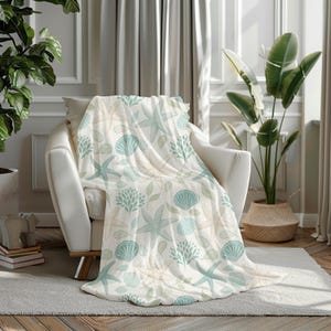 Puede incluir: Una manta blanca con un estampado de temática marina, sobre un sillón blanco. La manta presenta estrellas de mar, conchas marinas y follaje en tonos azules, verdes y beige. La manta es de un material suave.