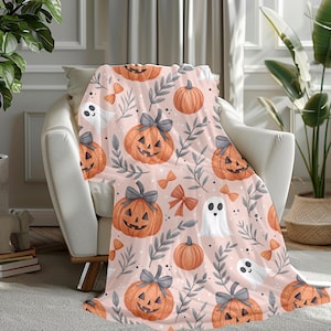 May include: A soft throw blanket with a light pink background featuring a repeating pattern of orange jack-o'-lanterns with grey bows, white ghosts, orange bows, and grey leafy branches. The design is consistent across the entire fabric surface.