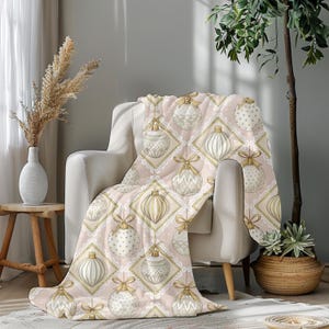 May include: A pink throw blanket featuring a repeating pattern of cream and gold Christmas baubles. The baubles are set within square frames with a lace edge and gold bows. The blanket is draped over a white armchair.