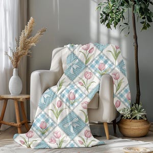May include: A soft, quilted throw blanket draped over a white armchair. The blanket features a repeating pattern of pink tulips, light blue gingham squares, and white bows with polka dots. The blanket is a cozy home decor item.