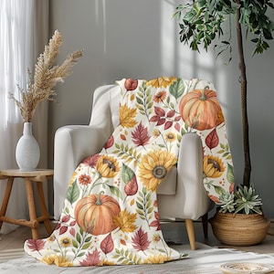 May include: A cream-colored throw blanket draped over a chair, featuring a watercolor design of pumpkins, sunflowers, and autumn leaves in shades of orange, yellow, and red. The blanket adds a cozy, seasonal touch to the room.