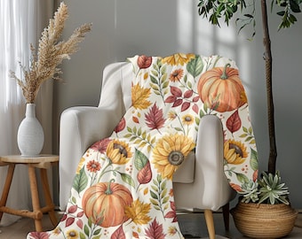 May include: A cream-colored throw blanket draped over a chair, featuring a watercolor design of pumpkins, sunflowers, and autumn leaves in shades of orange, yellow, and red. The blanket adds a cozy, seasonal touch to the room.