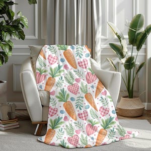 May include: A white blanket with a repeating pattern of orange carrots, pink gingham hearts, blue eggs, and green leaves. The blanket is draped over a white armchair in a well-lit room. The blanket is made of a soft material.