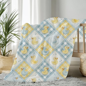 Pode incluir: Um cobertor de bebê com um padrão repetido de ilustrações de patos amarelos e azuis em um fundo xadrez branco, amarelo e azul. O cobertor está sobre um berço de madeira.