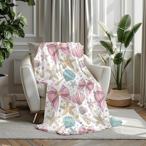 May include: A white throw blanket featuring a pattern of pink bows, seashells, starfish, and hearts. The blanket is draped over a white armchair, adding a decorative touch to a living room or bedroom.