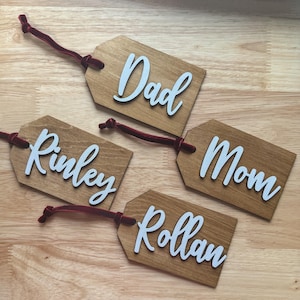 May include: Four wooden gift tags with white lettering and red velvet ribbon. The tags read "Dad", "Mom", "Rinley", and "Rollan". The tags are a light brown color and are arranged on a wooden surface.