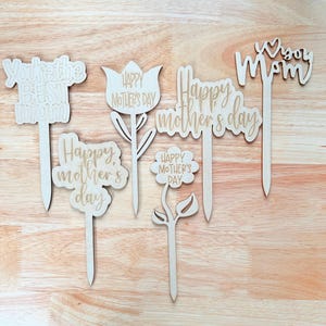 May include: Six wooden flower-shaped cake toppers with different Mother's Day messages. The messages include "You're the Best Mom", "Happy Mother's Day", "Happy Mother's Day", "Happy Mother's Day", "Happy Mother's Day", and "Happy Mom".