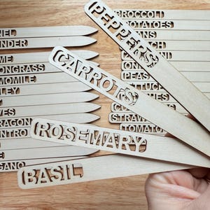 Puede incluir: Un conjunto de marcadores de plantas de madera con varios nombres de plantas recortados, incluyendo "Pimientos", "Zanahorias", "Romero" y "Albahaca".