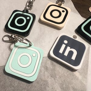 May include: Four keychains with different social media logos. The logos are Instagram, LinkedIn, and two different colored Instagram logos. The keychains are made of a white plastic with black and white logos.