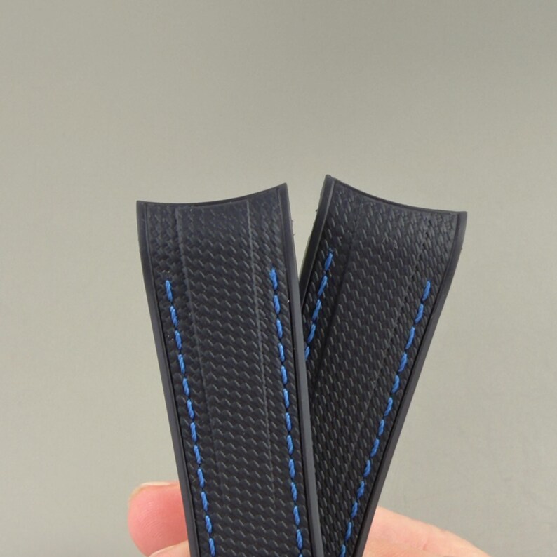 Puede incluir: Dos correas de reloj negras con un patr&oacute;n texturizado de fibra de carbono y costuras azules vibrantes en los bordes. Las correas tienen un dise&ntilde;o de extremo curvo destinado a encajar al ras contra la caja del reloj, con una estructura flexible.