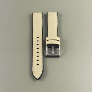 May include: A pair of watch straps with a light beige exterior and dark blue edges. The straps have a silver-colored buckle and a series of holes for adjustment. The watch straps are new.