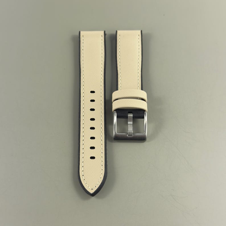 May include: A pair of watch straps with a light beige exterior and dark blue edges. The straps have a silver-colored buckle and a series of holes for adjustment. The watch straps are new.