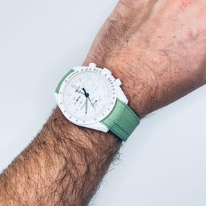 May include: A white wristwatch with a green strap. The watch face has a tachymeter scale and a chronograph function. The watch is on a person's wrist.