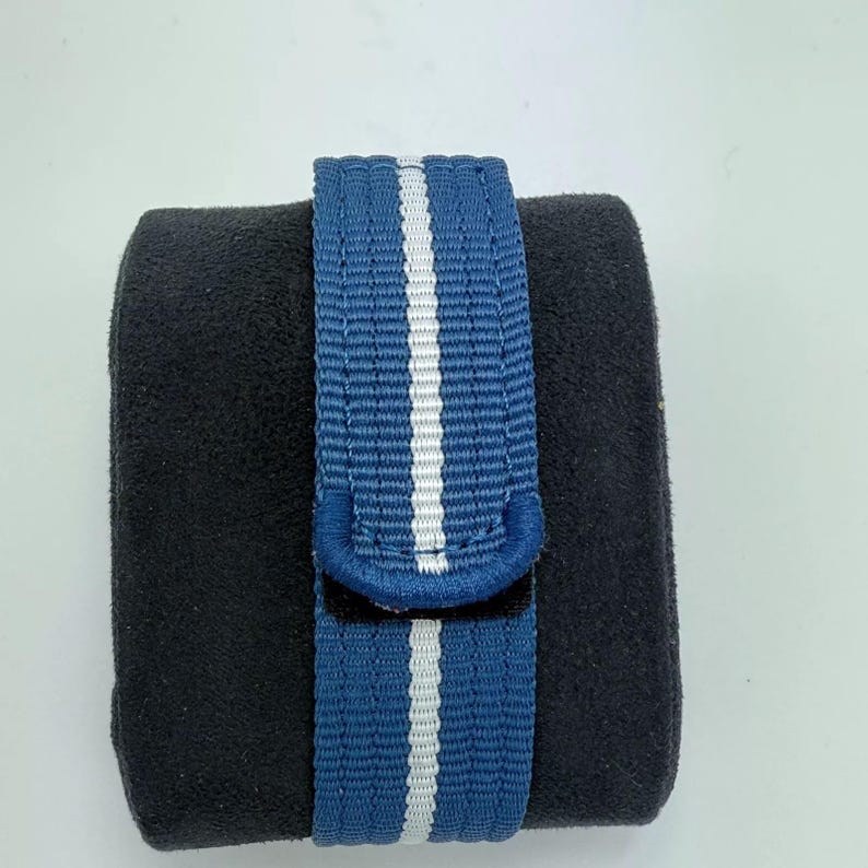 Puede incluir: Una correa de reloj azul que presenta una &uacute;nica franja blanca vertical que recorre el centro. La correa est&aacute; hecha de un material de tela tejida y est&aacute; envuelta alrededor de un coj&iacute;n de presentaci&oacute;n negro, rectangular y de textura aterciopelada.