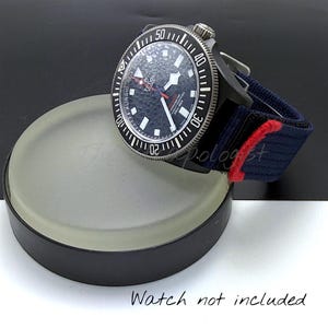 Könnte beinhalten: Eine schwarze und silberne Armbanduhr mit dunkelblauem Zifferblatt und schwarzer drehbarer Lünette. Die Uhr hat ein dunkelblaues und rot gestreiftes Armband. Die Uhr wird auf einem Ständer aus mattiertem Glas mit schwarzem Sockel präsentiert.