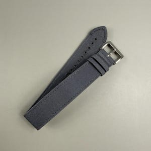 Puede incluir: Una correa de reloj de color gris oscuro hecha de un material de tela texturizada. La correa presenta costuras tonales a lo largo de los bordes, una serie de agujeros perforados para el ajuste y una hebilla de metal plateado pulido con trabillas.