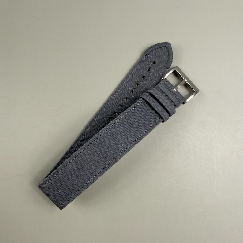 Puede incluir: Una correa de reloj de color gris oscuro hecha de un material de tela texturizada. La correa presenta costuras tonales a lo largo de los bordes, una serie de agujeros perforados para el ajuste y una hebilla de metal plateado pulido con trabillas.