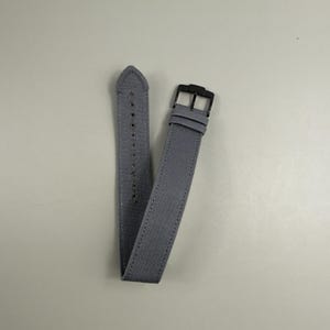 Puede incluir: Una correa de reloj de tela gris con un tejido texturizado, costuras tonales a lo largo de los bordes y una hebilla de metal negro. La correa incluye una serie de orificios perforados para el ajuste y dos trabillas de tela a juego.