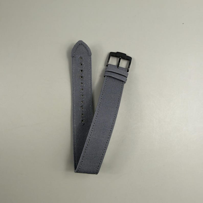 Puede incluir: Una correa de reloj de tela gris con un tejido texturizado, costuras tonales a lo largo de los bordes y una hebilla de metal negro. La correa incluye una serie de orificios perforados para el ajuste y dos trabillas de tela a juego.