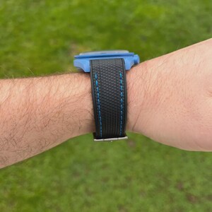 Puede incluir: Un reloj de pulsera con una caja azul brillante y una correa negra texturizada con costuras azules. El reloj se muestra desde una vista de perfil lateral, destacando el contraste entre la carcasa azul y el material oscuro y estampado.