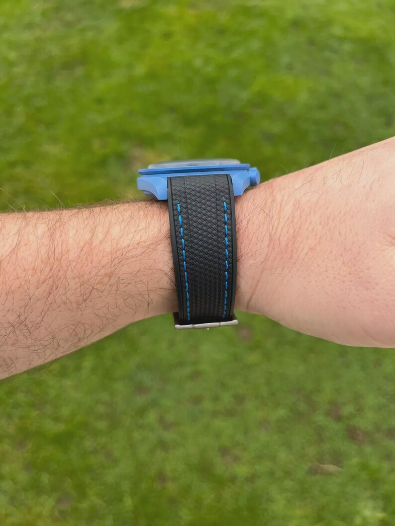 Puede incluir: Un reloj de pulsera con una caja azul brillante y una correa negra texturizada con costuras azules. El reloj se muestra desde una vista de perfil lateral, destacando el contraste entre la carcasa azul y el material oscuro y estampado.