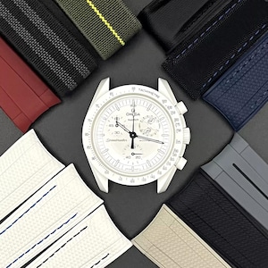 Może przedstawiać: Biały zegarek chronograf z czarną ramką tachymetru i białą tarczą. Zegarek jest otoczony r&oacute;żnymi paskami do zegark&oacute;w w r&oacute;żnych kolorach i materiałach, w tym czarnym, szarym, czerwonym, zielonym i niebieskim.