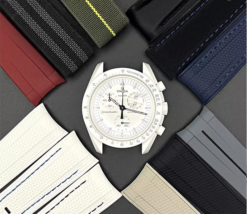 Może przedstawiać: Biały zegarek chronograf z czarną ramką tachymetru i białą tarczą. Zegarek jest otoczony r&oacute;żnymi paskami do zegark&oacute;w w r&oacute;żnych kolorach i materiałach, w tym czarnym, szarym, czerwonym, zielonym i niebieskim.