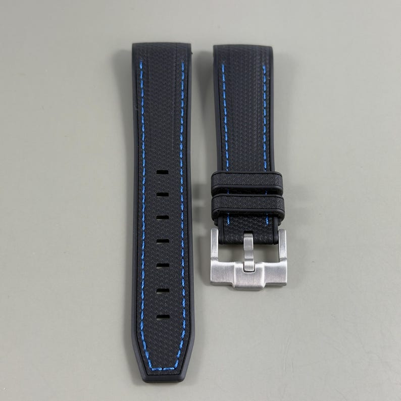 Puede incluir: Una correa de reloj negra con superficie texturizada y costuras azules. La correa consta de dos piezas, una con siete orificios de ajuste rectangulares y la otra con dos trabillas negras y una hebilla de metal plateado cepillado.