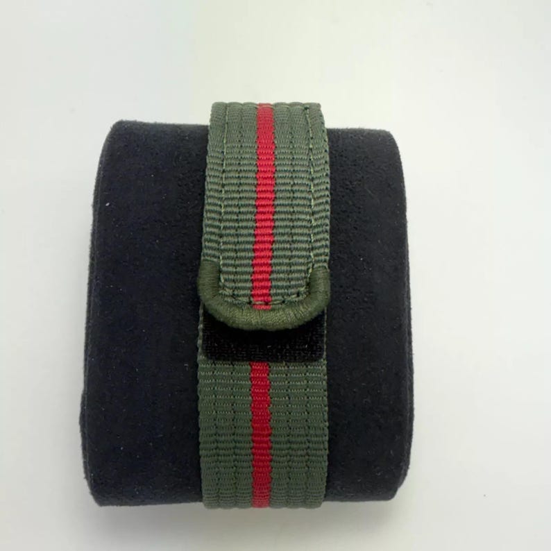 Puede incluir: Una correa de reloj de nailon verde oliva oscuro con una franja vertical roja en el centro. La correa est&aacute; envuelta alrededor de un coj&iacute;n de exhibici&oacute;n de textura aterciopelada negra, mostrando una construcci&oacute;n de tela tejida con bordes cosidos.