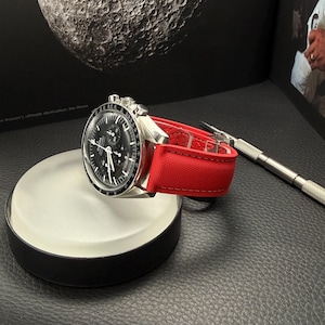 Puede incluir: Un reloj de pulsera plateado con esfera negra y correa roja descansa sobre un expositor blanco y negro. El reloj tiene una esfera detallada y un bisel texturizado. Una peque&ntilde;a herramienta plateada est&aacute; cerca.