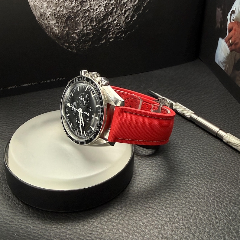 Puede incluir: Un reloj de pulsera plateado con esfera negra y correa roja descansa sobre un expositor blanco y negro. El reloj tiene una esfera detallada y un bisel texturizado. Una peque&ntilde;a herramienta plateada est&aacute; cerca.