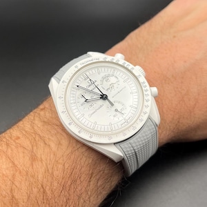 May include: A white wristwatch with a white strap and a tachymeter scale around the bezel. The watch face has a chronograph function and the text "Speedmaster" is printed on the face.
