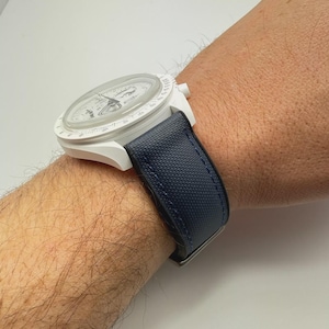 以下が含まれることがあります： 青いキャンバスストラップが付いた白いクロノグラフ腕時計。時計の文字盤は白地に黒の目盛りと針があります。