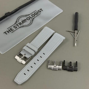 May include: Light gray watch strap with white stitching, a silver buckle, and a black and silver clasp. The strap has a textured pattern. The text "THE STRAPOLOGIST" is printed on a clear pouch. A small screwdriver is also included.