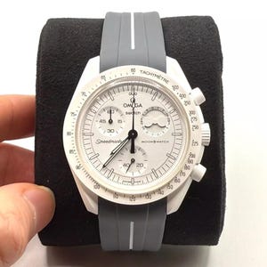 Może przedstawiać: Biało-szary zegarek na rękę z chronografem i tachymetrem. Tarcza zegarka ma nadrukowane słowa "Speedmaster" i "Moonswatch". Zegarek ma szary silikonowy pasek.