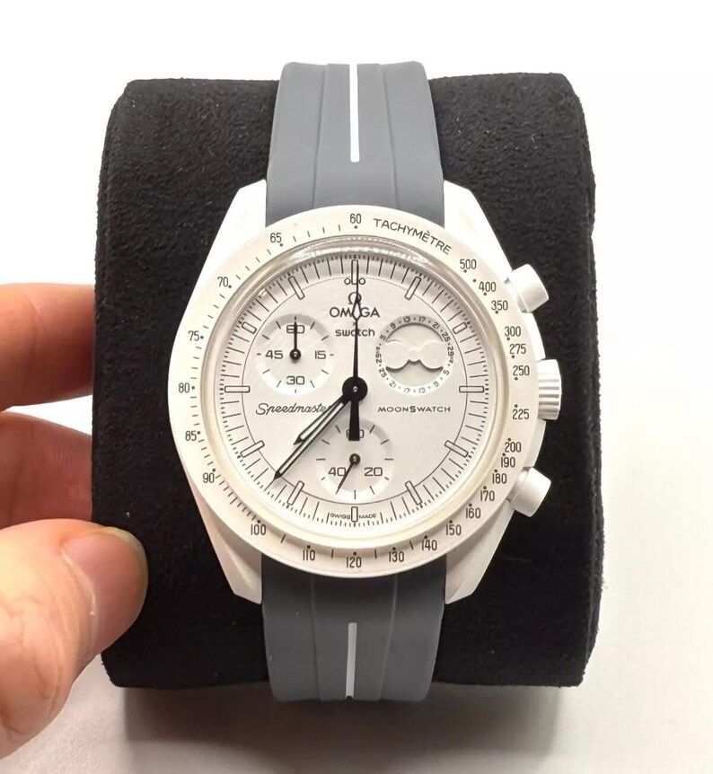 Może przedstawiać: Biało-szary zegarek na rękę z chronografem i tachymetrem. Tarcza zegarka ma nadrukowane słowa "Speedmaster" i "Moonswatch". Zegarek ma szary silikonowy pasek.