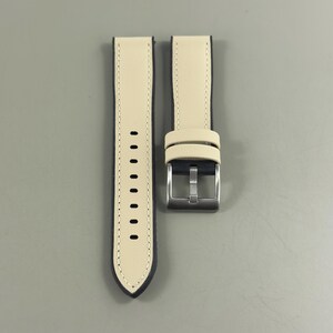 May include: A pair of watch straps with a cream-colored top and a dark blue underside. The straps have a stainless steel buckle and a series of holes for adjustment. The stitching is visible along the edges.
